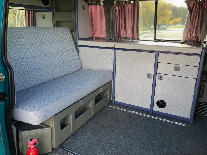 Inside this RV rental: a patterned bench seat, integrated kitchenette with storage, and dark window curtains for comfort.. Volkswagen T4 Westfalia Coach 1992