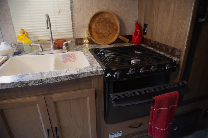 Compact RV rental kitchen: light wood cabinets, granite-look counter, sink, black gas stove/oven with red towel.. Forest River Coachmen Prism 2018