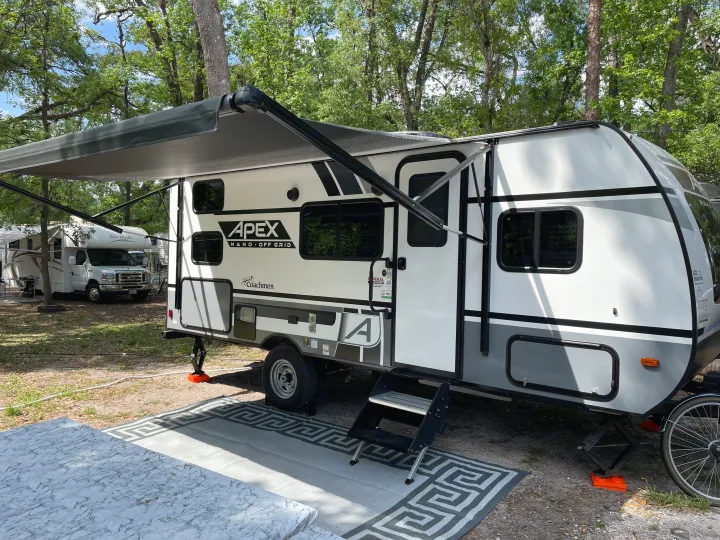 A modern white and gray Apex Nano travel trailer, perfect for an RV rental, is set up with an awning at a wooded campsite.. Coachmen Apex Nano 2022