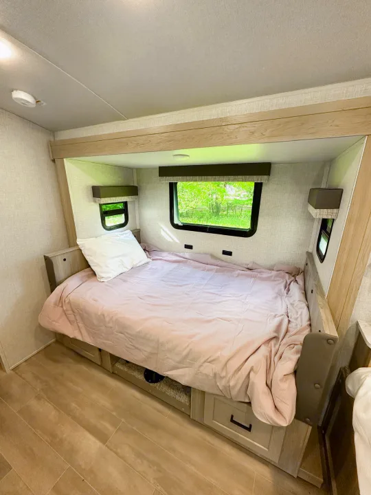 Cozy RV rental bedroom with a light pink bed, under-bed storage, and scenic nature views through the windows.. Forest River Flagstaff E-Pro 2023