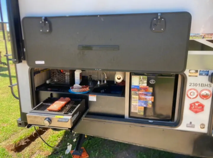 This RV rental's outdoor kitchen features a grill cooking hot dogs, a sink, and a mini-fridge. Perfect for camping!. Winnebago Minnie 2022