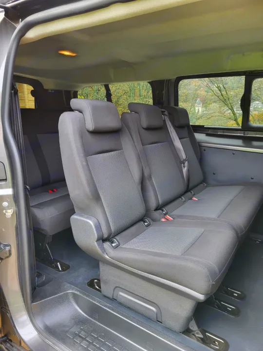 Comfortable, black passenger seating in this multi-row RV rental van. View of trees and a distant building through side windows.. Peugeot Expert combi long 2022