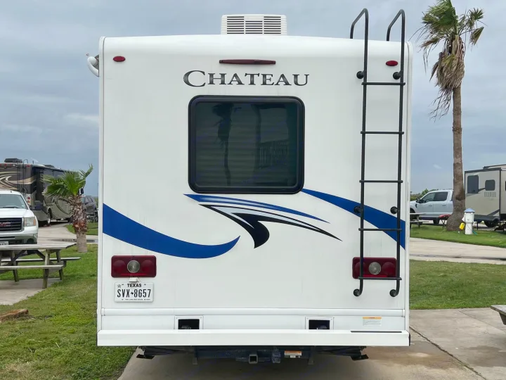 Rear view of a white Chateau RV rental, license plate SVX-8657, parked at an RV park with palm trees on a cloudy day.. Thor Chateau 2020