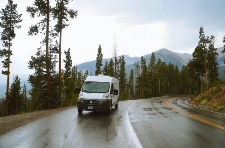White camper van (RV rental) on a wet, winding mountain road. Tall pines and distant peaks create a scenic travel vista.. Other Sprinter Van 2017