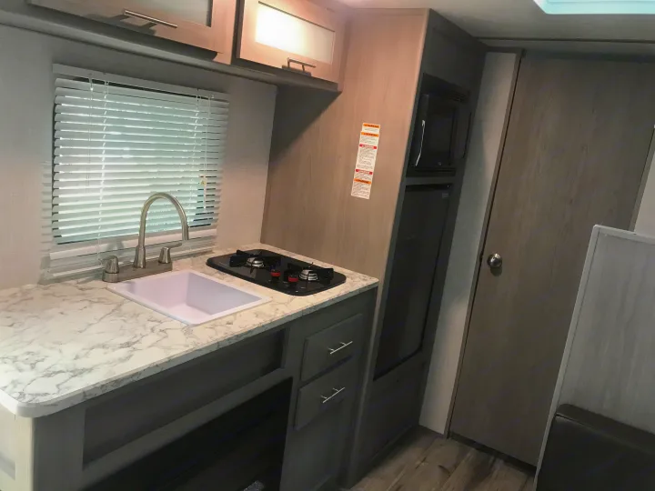 Compact kitchen in an RV rental featuring a white sink, two-burner stove, overhead cabinets, microwave, and fridge.. Dutchmen Coleman Lantern 2023