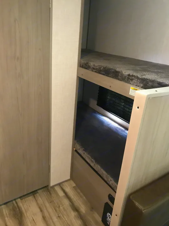 RV rental bunk beds (top bunk rated 300 lbs max) with textured grey mattresses, a privacy shade, and a light wood door.. Dutchmen Coleman Lantern 2023