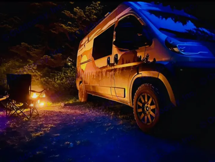 Cozy RV rental night: Camper van with scenic decal, glowing campfire, and chair. Serene camping scene.. Dodge Ram 1500 2020