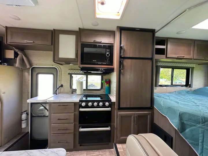 Modern RV rental interior showing a full kitchen with stove, microwave, and fridge, plus a cozy bed area.. Thor Coleman 2023