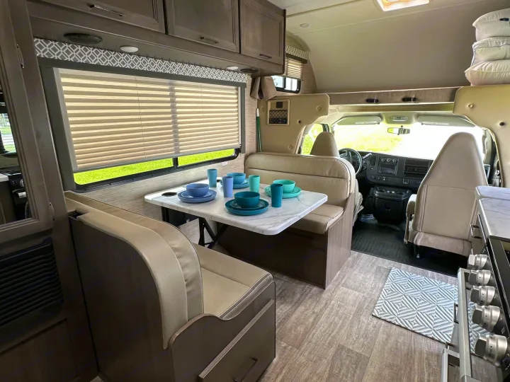 Bright RV rental interior showcasing a kitchen with stove, a dinette with colorful dishes, and the front cockpit area.. Thor Coleman 2023
