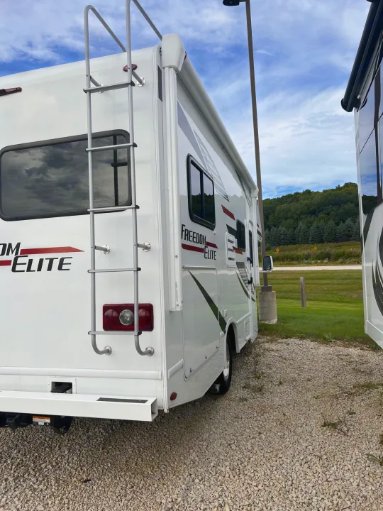 White Freedom Elite RV rental, a Class C motorhome, parked on a gravel lot with a ladder on its rear. Blue sky and trees visible.. Thor Freedom Elite 2020
