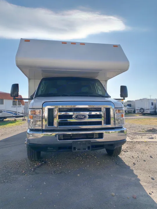 A white Ford Class C RV rental is seen from the front, parked on a gravel lot with other RVs nearby under a blue sky.. Thor Majestic 2017