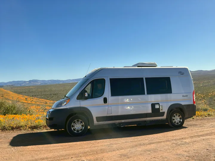 A silver camper van (RV rental) on a dirt road, amidst orange wildflowers with hills under a clear blue sky.. Carado Carado Class B 2018
