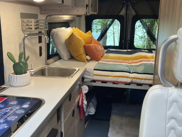 Cozy rv rental interior featuring a well-appointed kitchenette, bed with colorful pillows, and serene lake views from the back.. Winnebago SOLIS 59 PX 2023