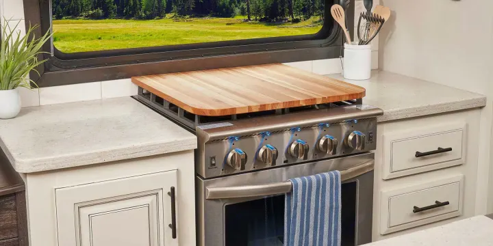 An RV rental kitchen, showcasing a stainless steel stove with a wooden cover, light countertops, and a window view of nature.. Jayco Pinnacle 2022