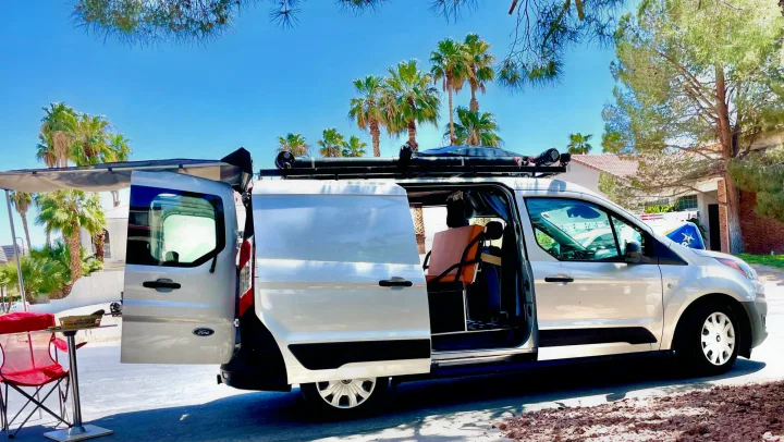Sunny day setup for an rv rental: a light van with open doors, an extended awning, & a red camping chair.. Ford transit connect 2022