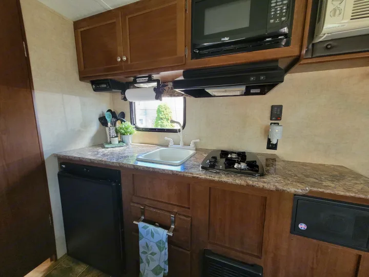 This RV rental kitchen features a sink, two-burner stove, microwave, and mini-fridge, with wood cabinets.. Keystone RV Hideout LHS 2016