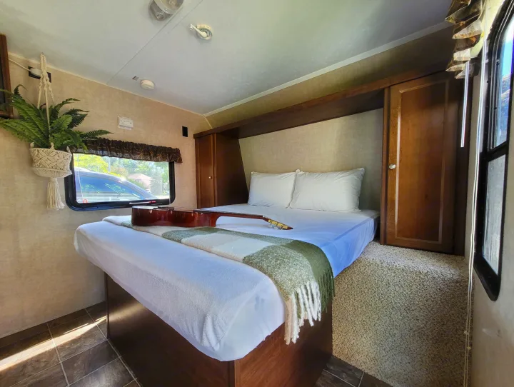 Spacious RV rental bedroom with a cozy bed, acoustic guitar, and hanging fern. Features warm wood cabinetry and natural light.. Keystone RV Hideout LHS 2016