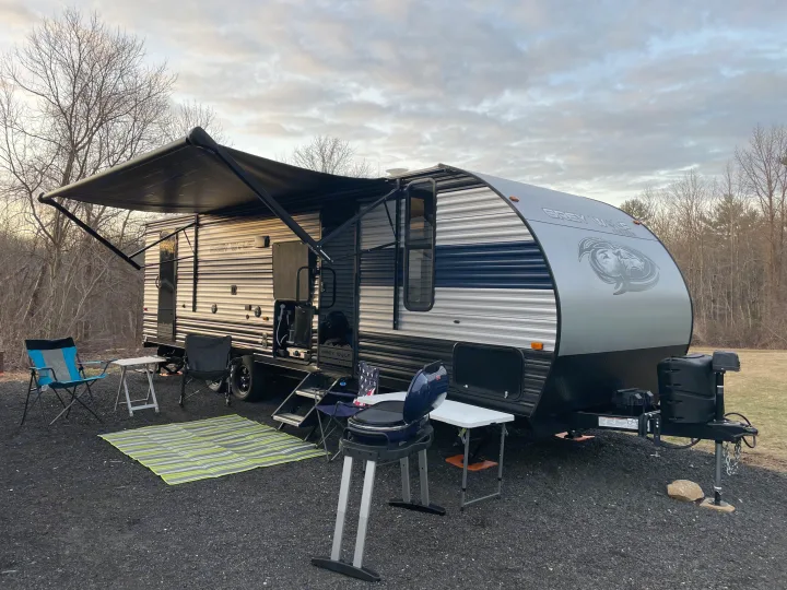 RV rental camping setup with an extended awning, chairs, grill, and outdoor mat on a gravel site.. Forest River Cherokee Grey Wolf 2022