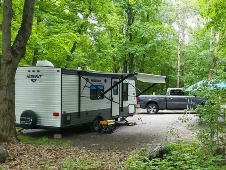 An RV rental travel trailer is parked at a wooded campsite with an extended awning, generator, and a pickup truck.. Keystone RV Hideout LHS 2016