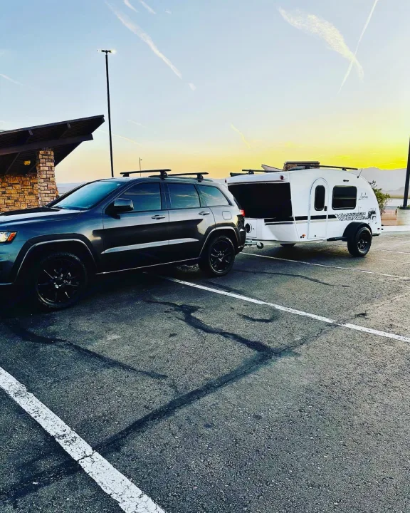 Dark SUV with roof racks hitches to a white teardrop RV rental, a Luna Rover, in a parking lot at sunset.. InTech RV InTech RV Trailer 2021