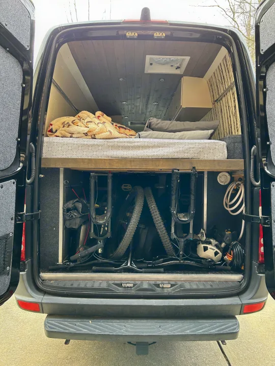 Rear view of a spacious custom RV rental camper van, revealing a comfortable bed above organized mountain bike storage.. Mercedes Sprinter 2012