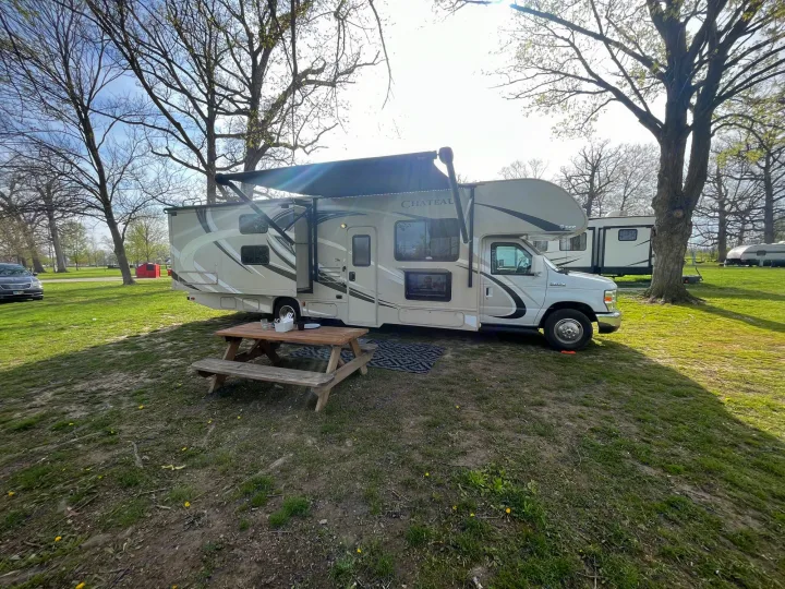 Class C RV rental with awning extended, parked on a grassy campground site with a picnic table. Sunny day, ideal for camping.. Thor Chateau 2017