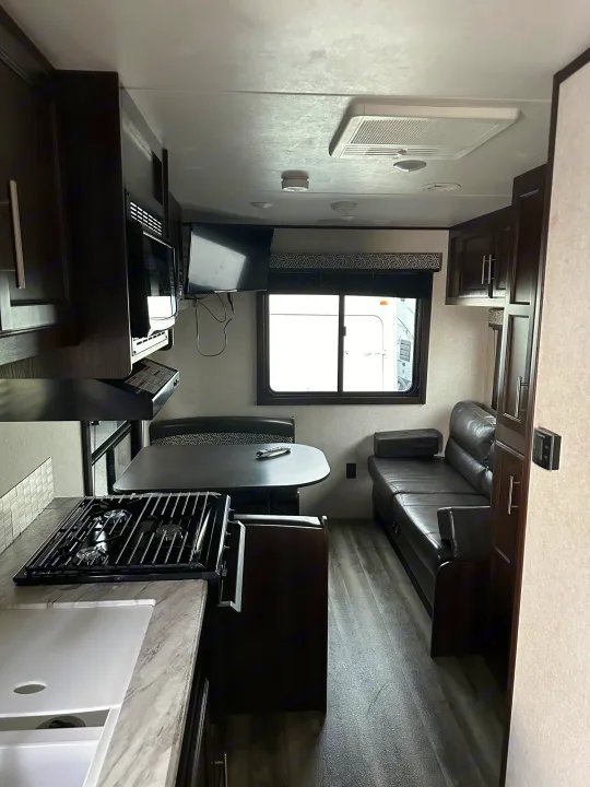 Modern RV rental interior: kitchen with stovetop, dining booth, and dark sofa. Features dark wood and light accents.. Jayco Flight 2020