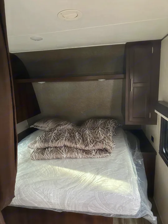 RV rental bedroom showing a bed with plastic-wrapped mattress, patterned bedding, and dark wood cabinetry.. Jayco Flight 2020