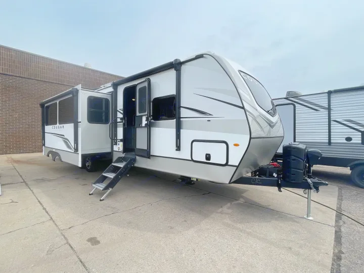 Modern white & gray travel trailer RV rental with extended slide-out, open door, and steps down, parked on concrete.. Keystone Cougar 2023