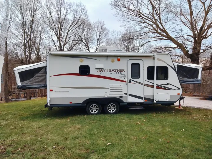 White Jay Feather Ultra Lite travel trailer RV rental, with pop-out beds extended, parked on green grass among bare trees.. Jayco Jay Feather Ultra Lite 2013