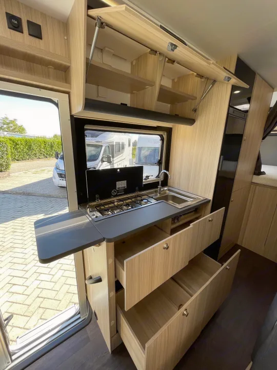 Inside an RV rental, a modern kitchen with open drawers, overhead storage, stove, sink, and an extendable counter. View of another RV outside.. SunLiving A 75 DP 23 2023