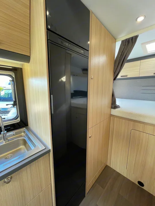 RV rental interior featuring a kitchen sink, tall black fridge, wooden cabinetry, and a cozy bed area.. SunLiving A 75 DP 23 2023