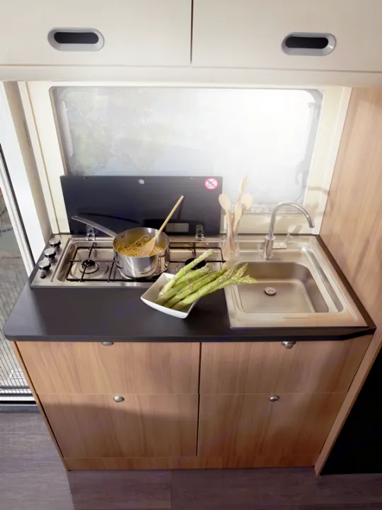 RV rental kitchen: compact stove, sink, wooden cabinets. Pasta cooks, fresh asparagus awaits on counter.. SunLiving A 75 DP 23 2023