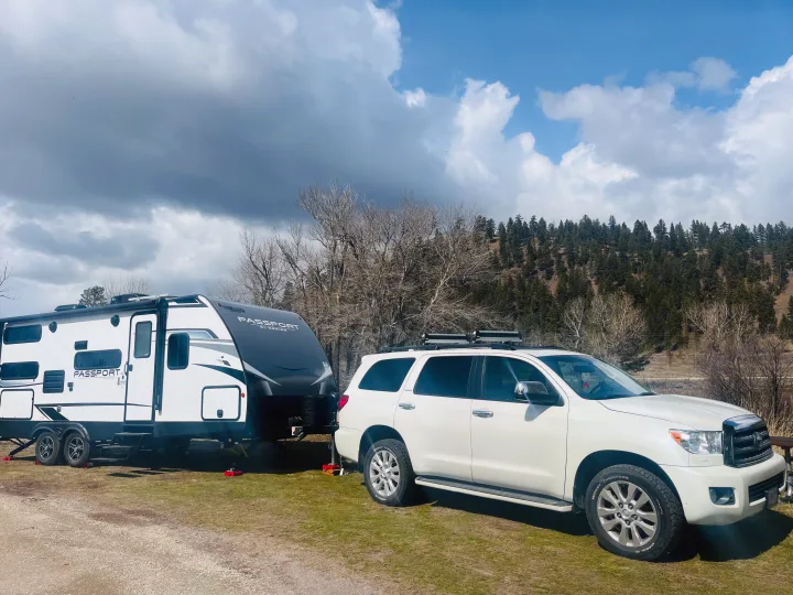 White SUV with a Keystone Passport **RV rental** at a scenic campground. Ready for adventure amidst trees and mountains under a cloudy sky.. Keystone RV Passport SL 2022