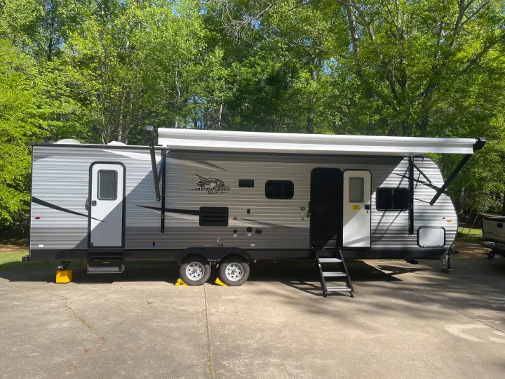 This gray and black Jayco Jay Flight SLX travel trailer, a great option for an RV rental, is parked on concrete with an awning extended, surrounded by green trees.. Jayco Jay Flight SLX 2021