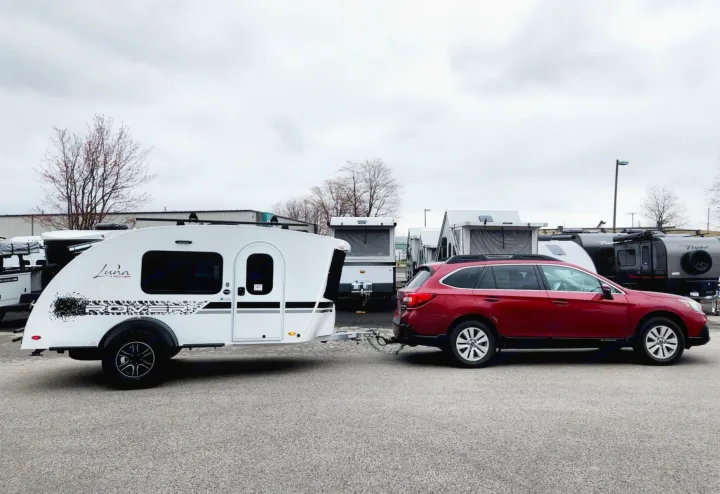 White teardrop RV rental hitched to a red Subaru Outback, ready for adventure. Pop-ups & other RVs in the background.. Intech Luna Rover 2023