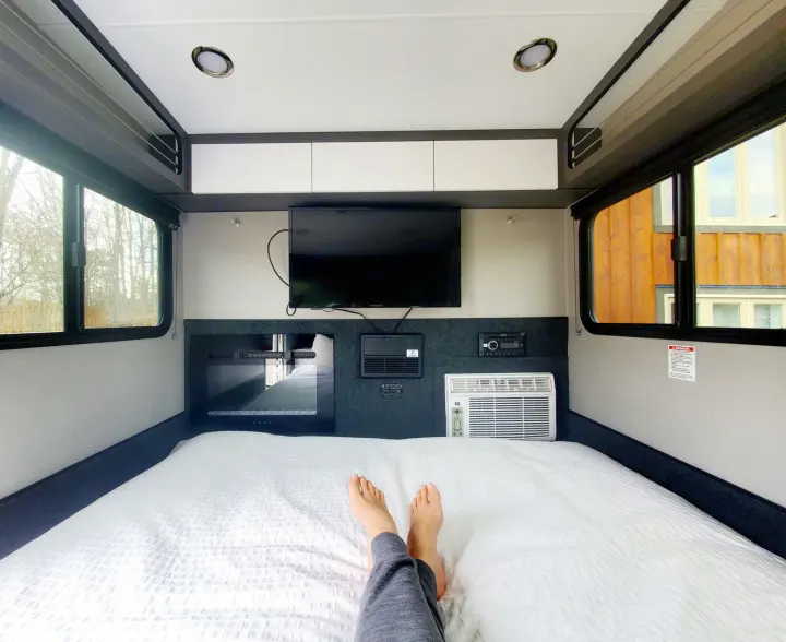 Cozy RV rental interior with a comfy bed, TV, and AC unit. A person relaxes with feet up, enjoying the view.. Intech Luna Rover 2023