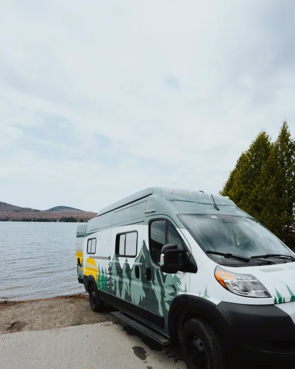 An rv rental camper van with mountain decals is parked by a scenic lake with hills, ready for adventure.. Winnebago Solis 2022