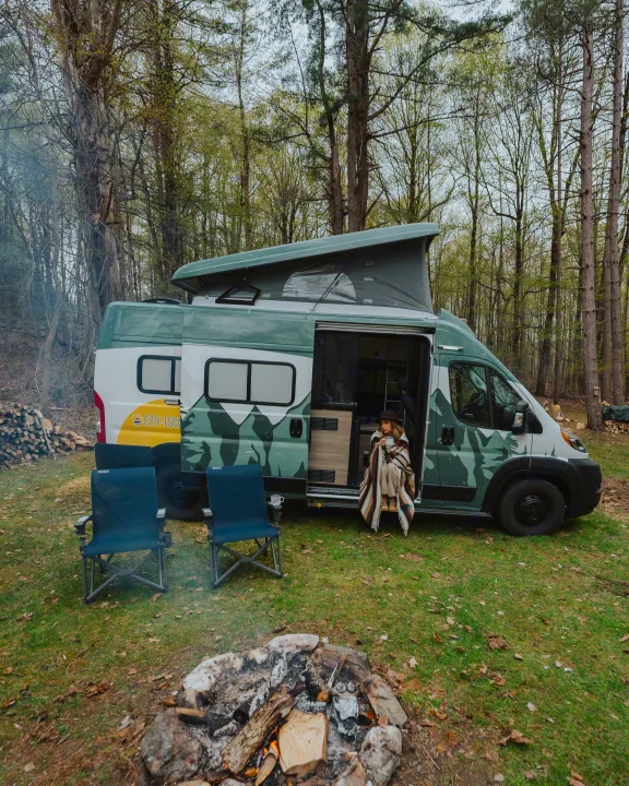 Cozy rv rental experience: a pop-top camper van with a person relaxing, chairs, and a crackling campfire in nature.. Winnebago Solis 2022