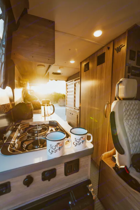 Warm sun streams into a cozy RV rental kitchen with a stove, French press, and "To Tour Shore" mugs.. Winnebago Solis 2022