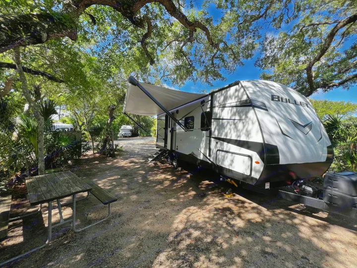 A white travel trailer RV rental, with its awning extended, is parked at a shaded campsite alongside a picnic table.. Keystone RV Bullet Ultra Lite 2022