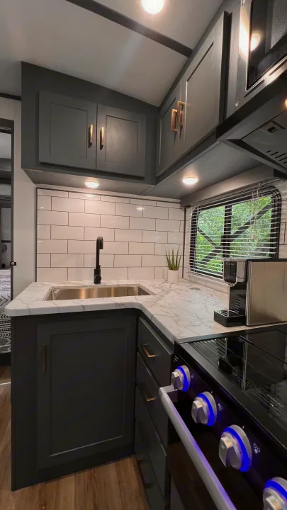 Modern RV rental kitchen with dark cabinets, marble counters, and white subway tile backsplash. Blue illuminated stove knobs add a sleek touch.. Keystone RV Bullet Ultra Lite 2022