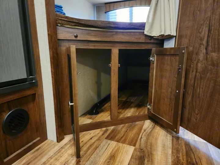 RV rental interior: open cabinets under a bunk bed, revealing storage space with a pipe, plus wood flooring and a vent.. Cruiser RV MPG Ultra Lite 2017