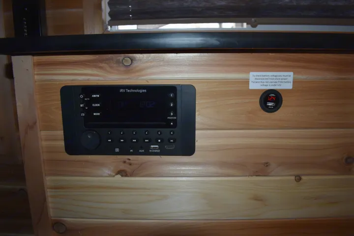 RV rental interior: iRV Technologies stereo, digital voltage display (12.4V), and battery info sticker on wood paneling.. Ice Castle Fish Houses Rv Edition Standard 2024