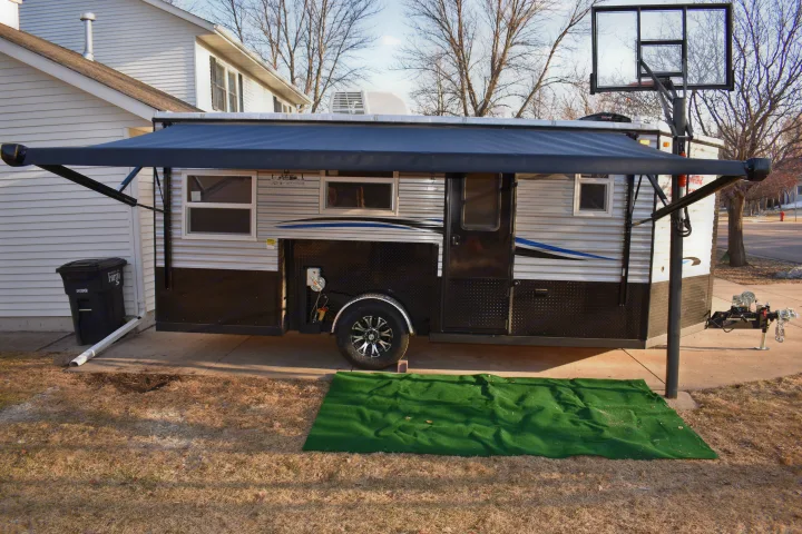 RV rental: A dark custom travel trailer with an extended awning and a green mat, parked in a driveway by a house.. Ice Castle Fish Houses Rv Edition Standard 2024