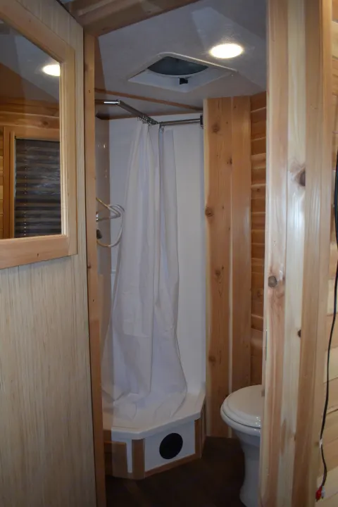 Inside an RV rental: a compact bathroom with a shower (white curtain) and toilet, all finished in natural wood.. Ice Castle Fish Houses Rv Edition Standard 2024
