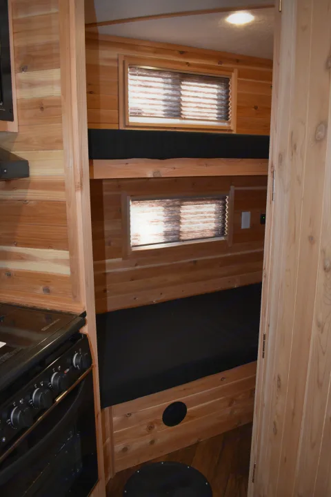 Inside an RV rental: wooden interior with stacked bunk beds, a compact kitchen stove, and windows for cozy adventures.. Ice Castle Fish Houses Rv Edition Standard 2024