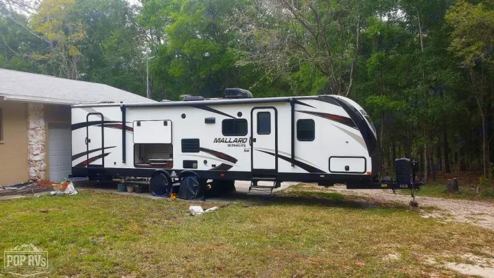White Mallard Ultralite RV rental, a modern travel trailer parked on grass next to a house and trees.. Heartland RVs Mallard 2019