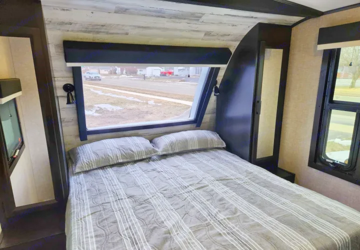 Experience comfort in this RV rental bedroom: cozy bed, panoramic window with street view, and integrated storage.. Jayco Whitehawk 2023