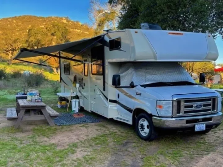 An RV rental parked at a tranquil campsite, complete with an extended awning and picnic area.. Coachmen Leprechaun 2016